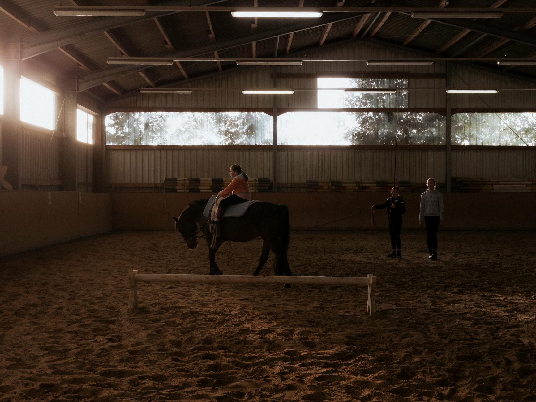 Lekcja jazdy konnej w hali, dziecko na koniu z asekuracją dorosłych